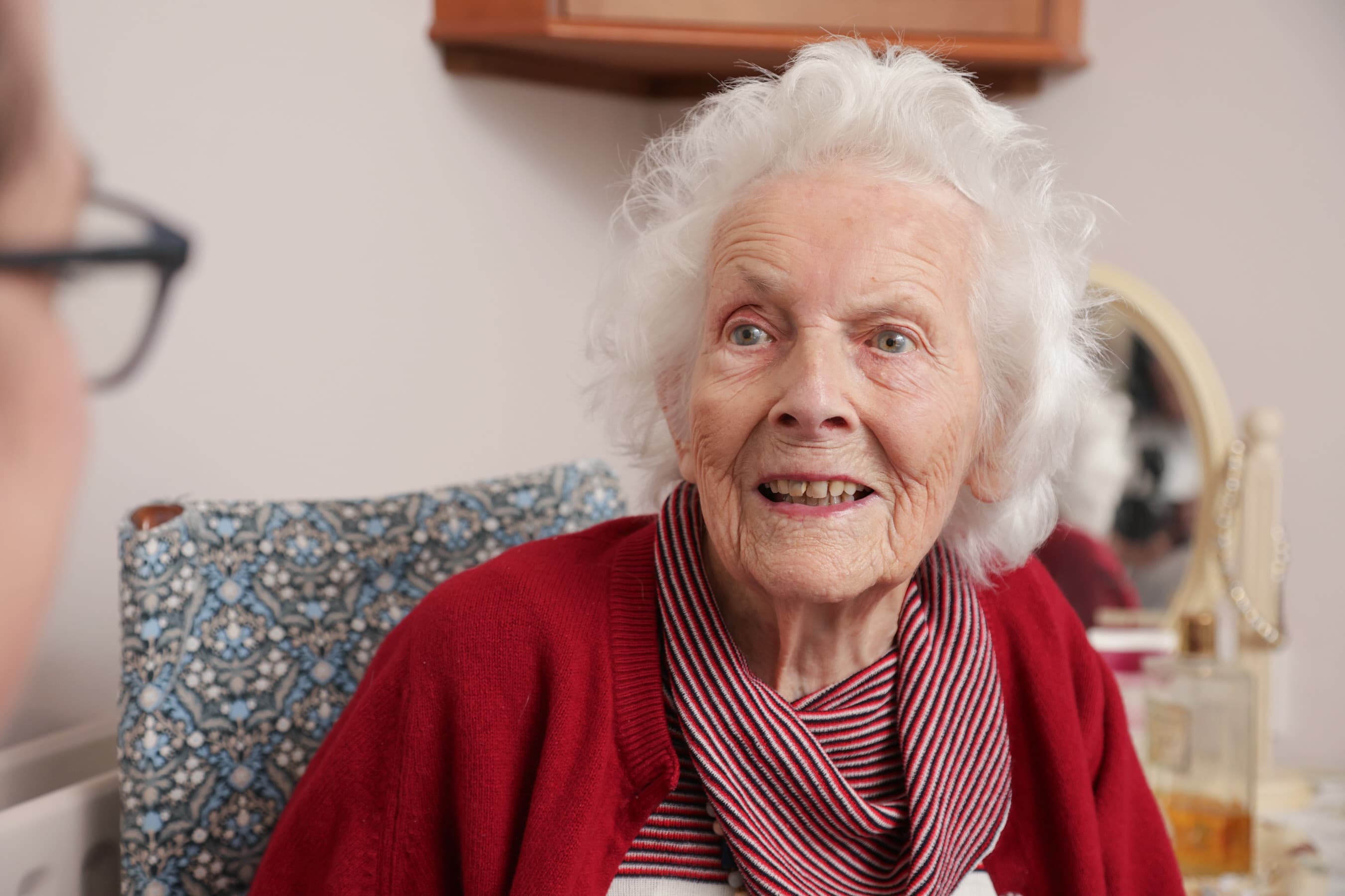 Elderly woman with white hair and red sweater smiling, sitting indoors and talking to someone. - Home Instead