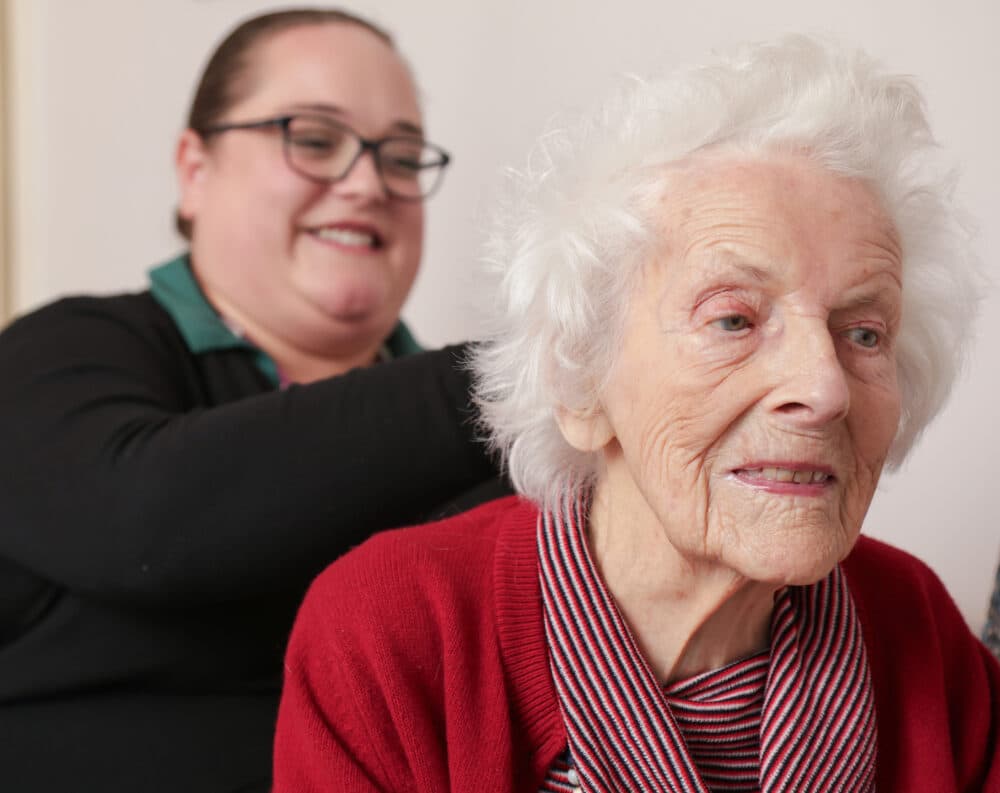 Smiling elderly woman with white hair and red sweater sits as a caregiver stands behind her, also smiling. - Home Instead