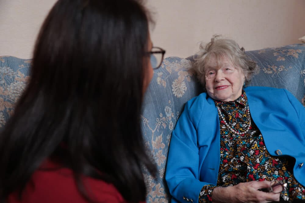 An elderly woman in a blue jacket smiles while talking to another person on a patterned couch. - Home Instead