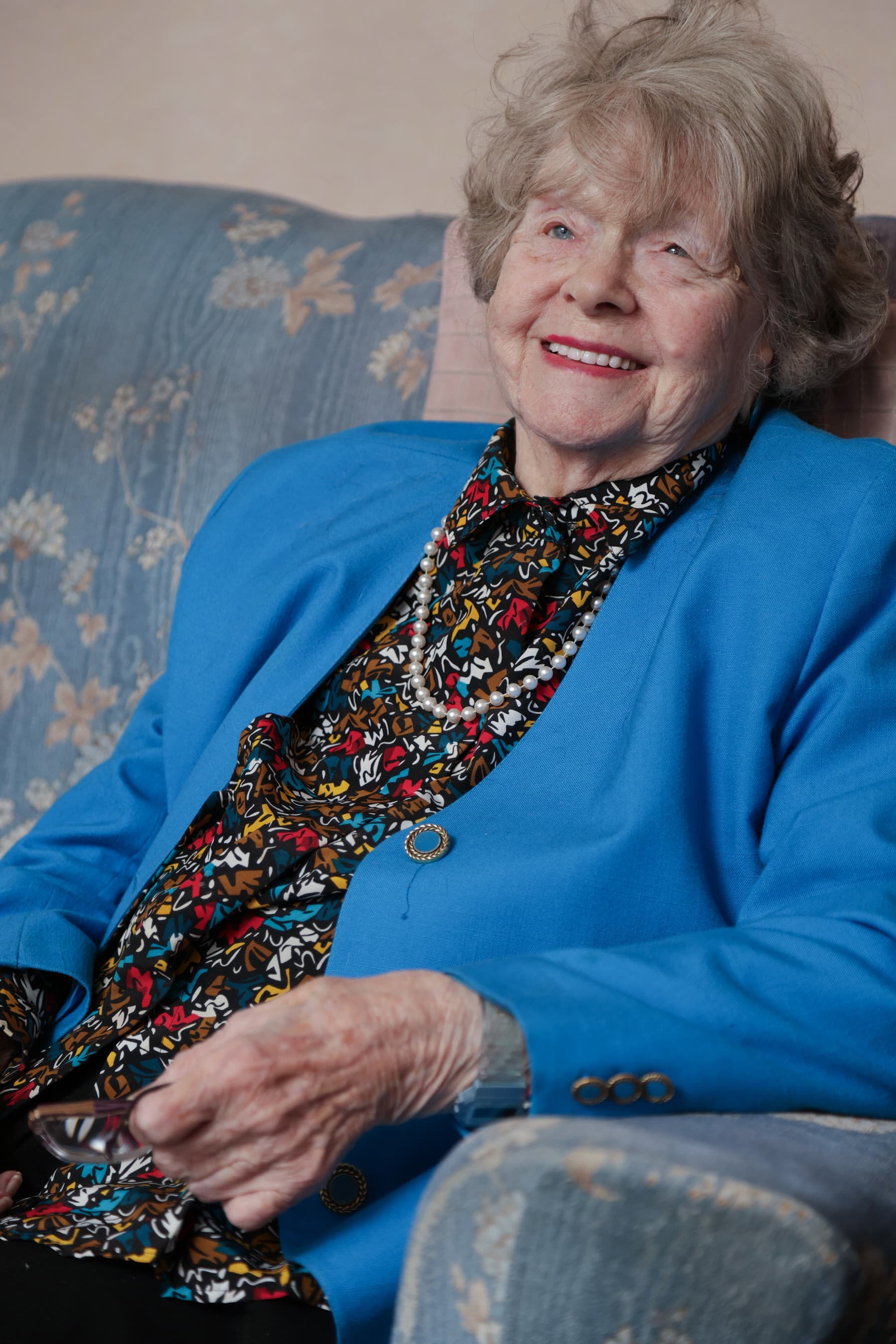 Smiling elderly woman in a blue blazer sits on a patterned couch, holding glasses in her hand. - Home Instead