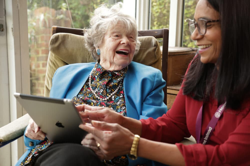 Two women smiling and using a tablet together while sitting indoors near a window. - Home Instead