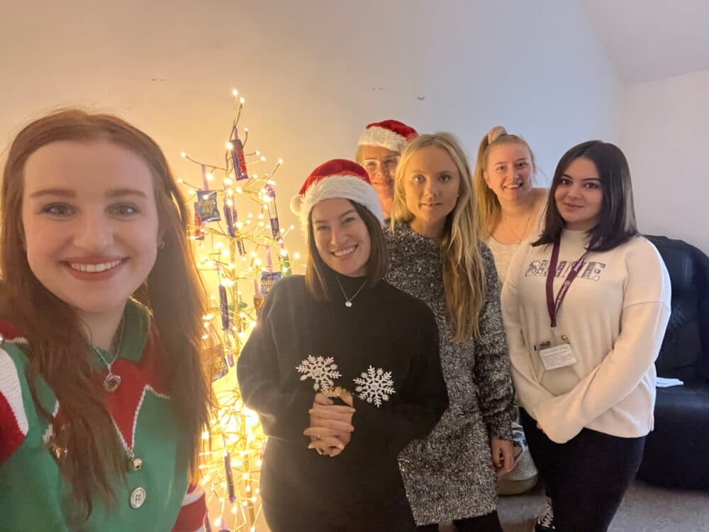 Six people pose and smile by a lit Christmas tree, some wearing festive hats and sweaters. - Home Instead
