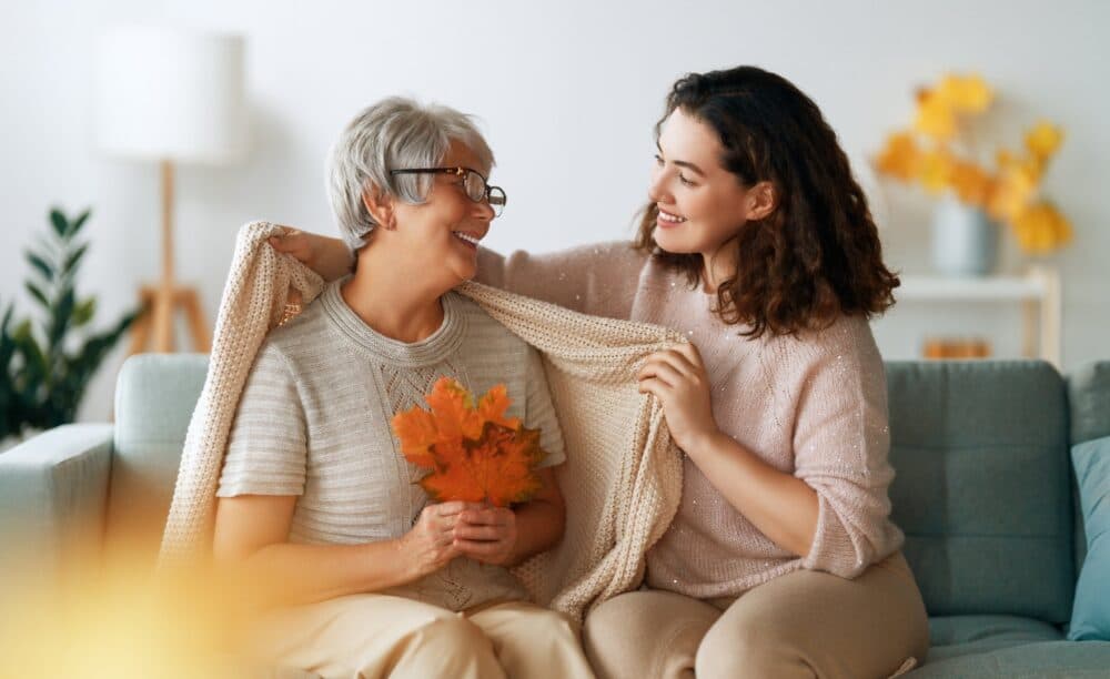 Smiling woman wraps a cozy shawl around an older woman holding autumn leaves on a couch. - Home Instead