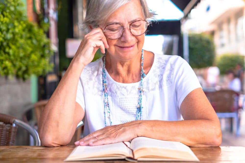 Smiling older woman with glasses reading a book at an outdoor café table. - Home Instead