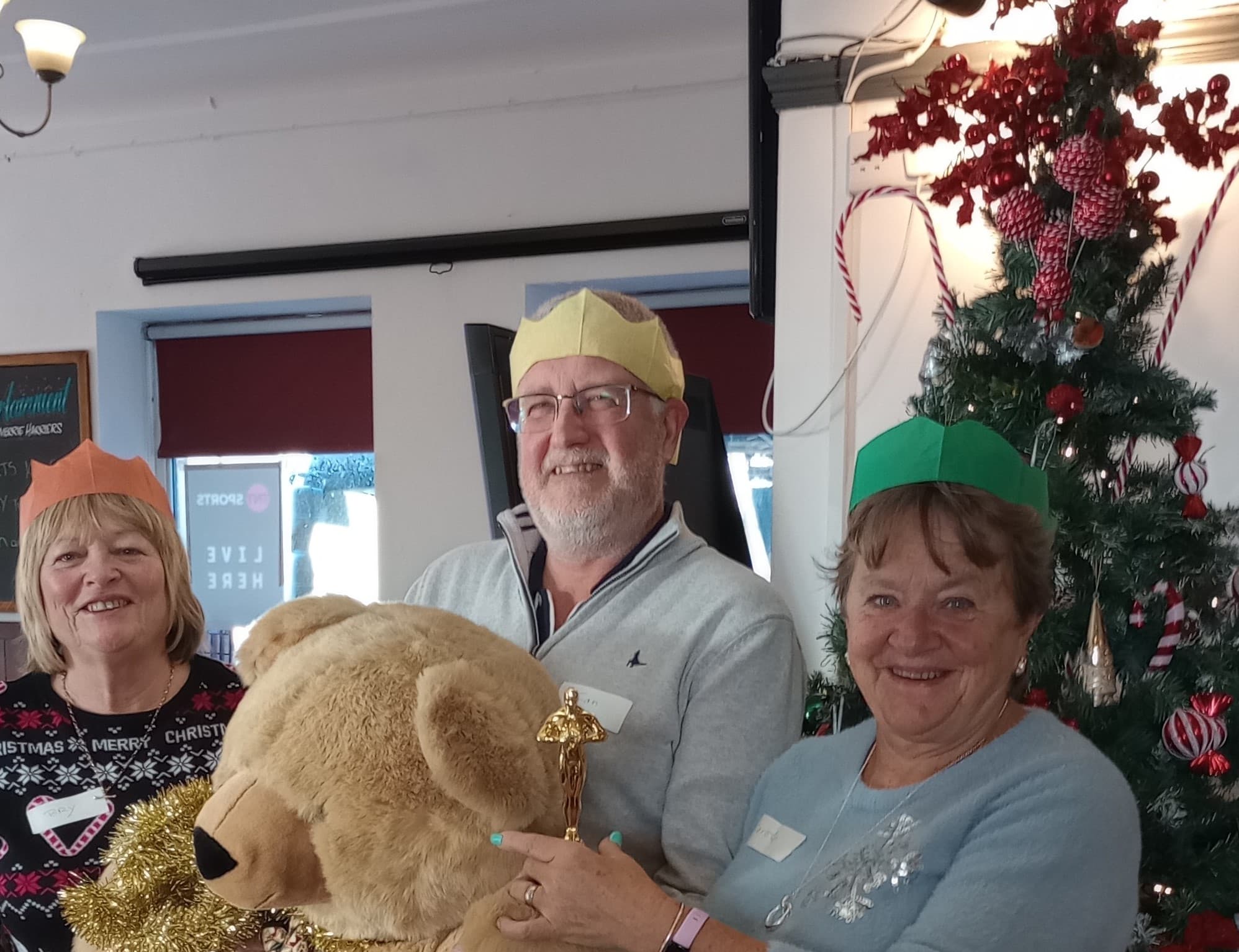 Three smiling adults in party hats stand by a Christmas tree, holding a large teddy bear and a gold trophy. - Home Instead