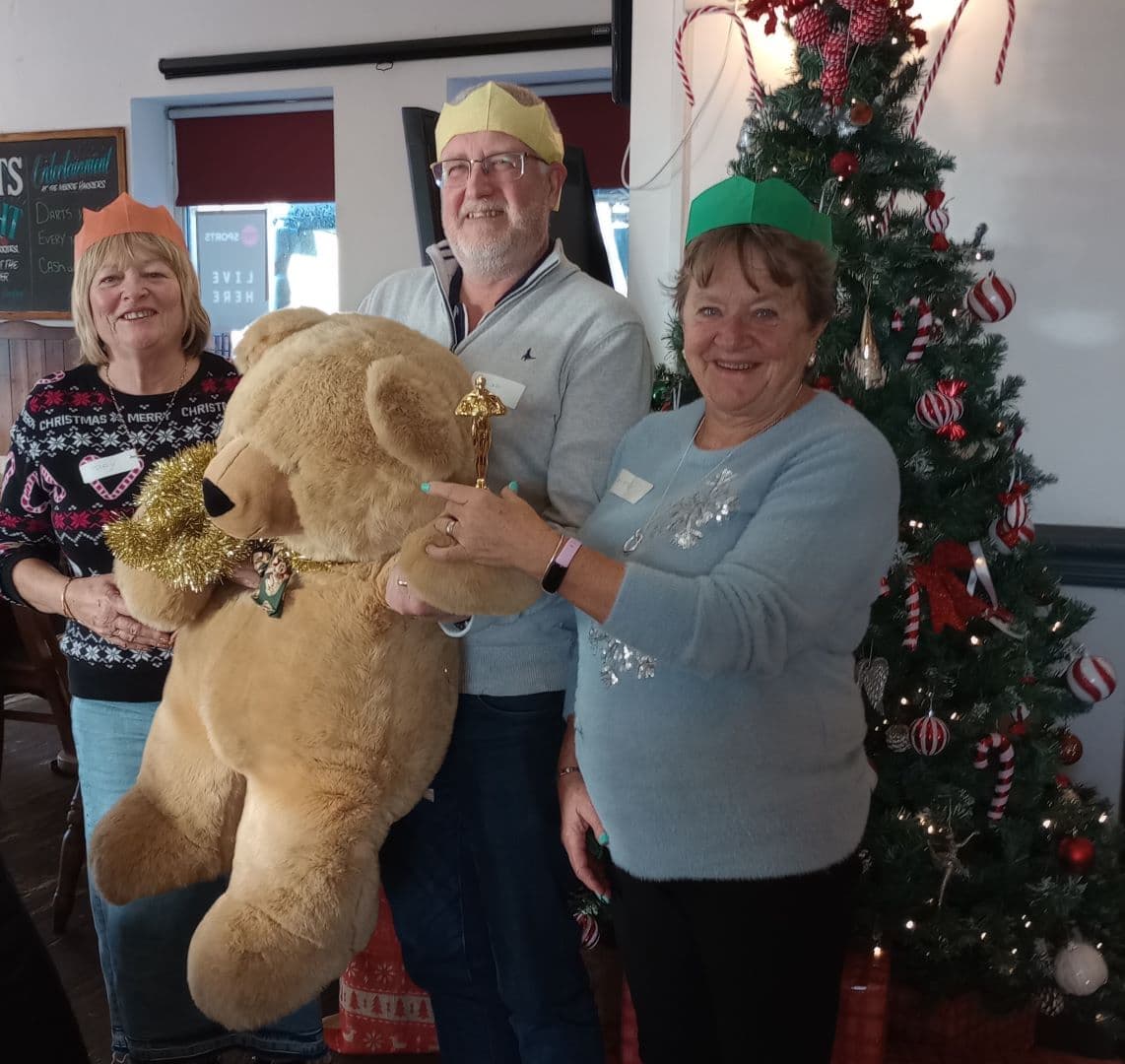 Three smiling adults in festive hats hold a large teddy bear beside a decorated Christmas tree. - Home Instead