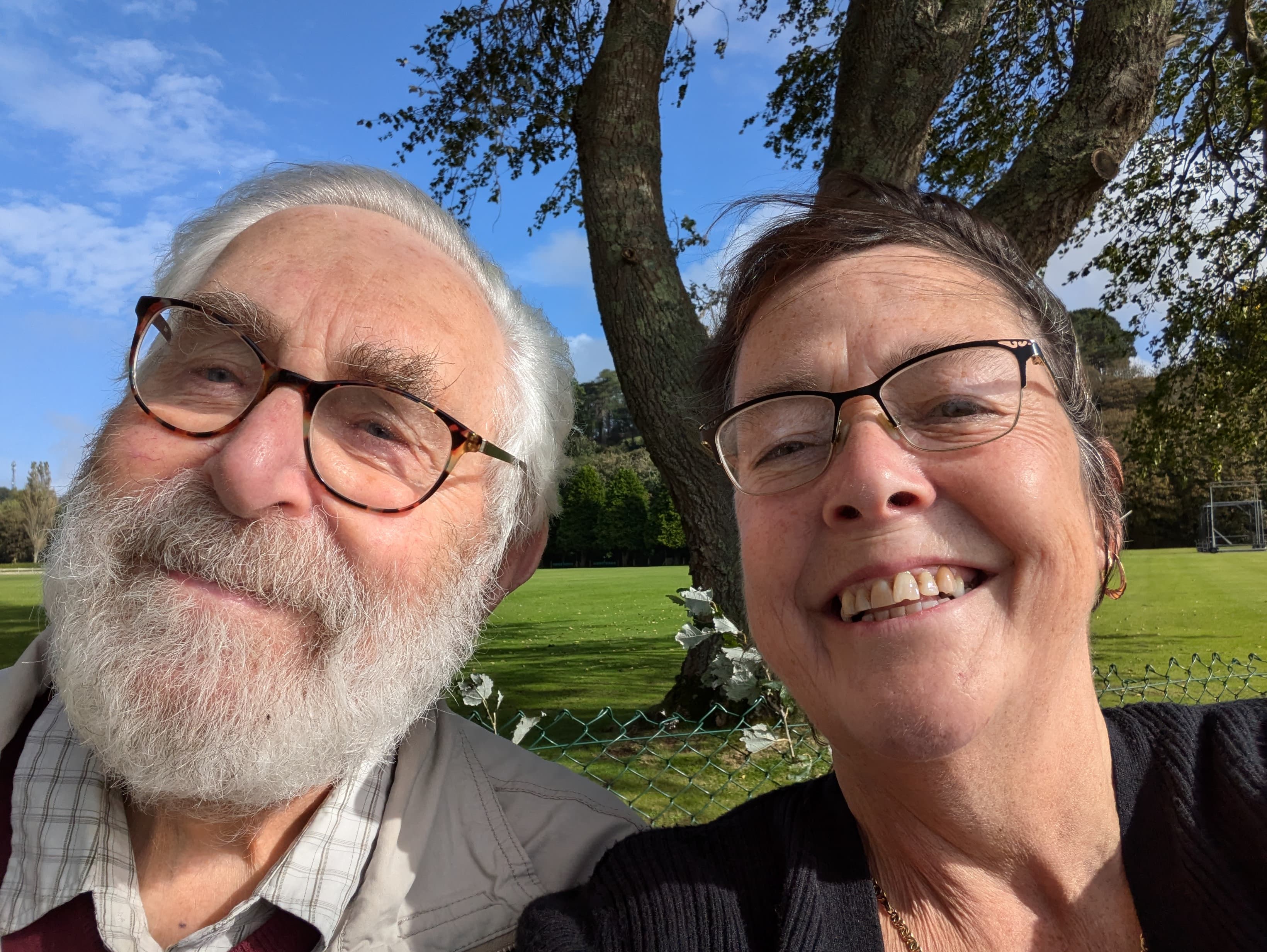 Smiling elderly man and woman with glasses pose for a selfie outdoors on a sunny day near a tree. - Home Instead