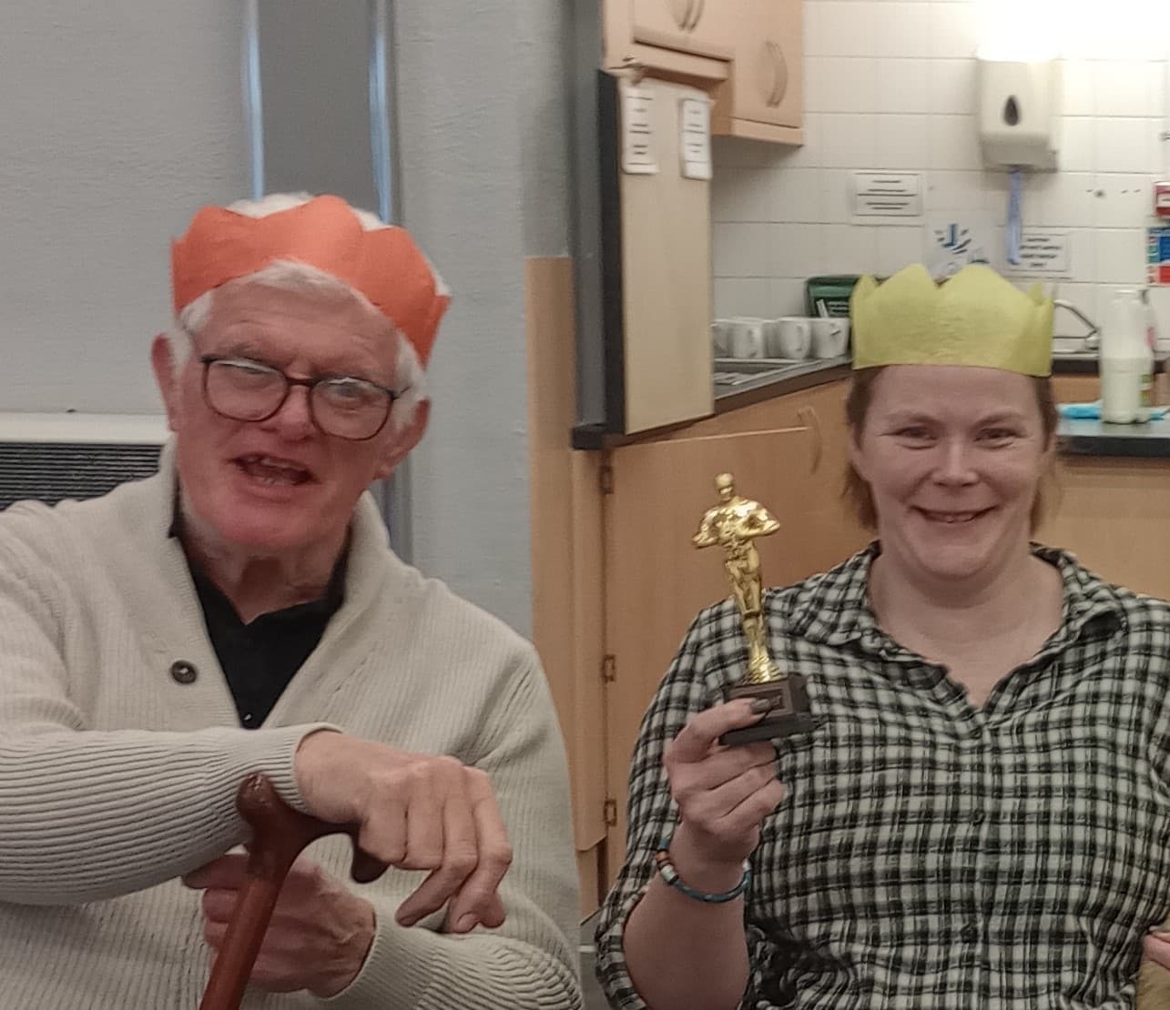 Two people wearing paper crowns, one holding a cane and the other holding a trophy, smiling indoors. - Home Instead