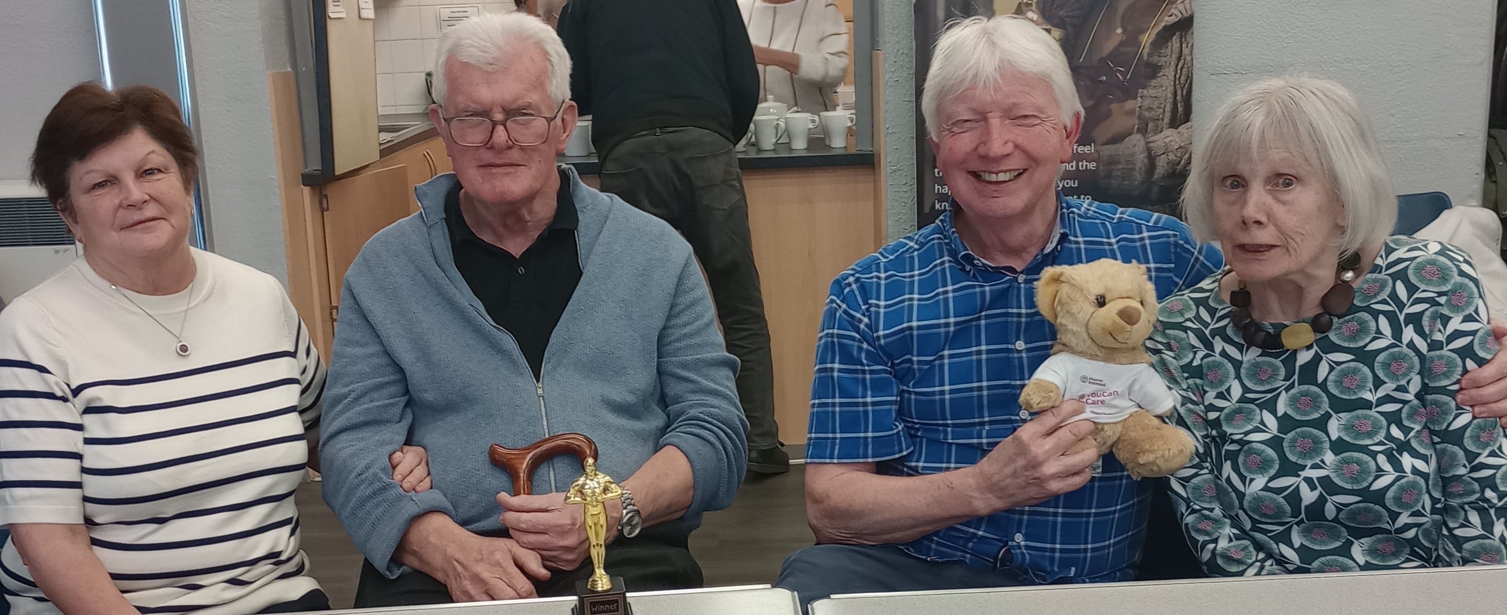 Four older adults sit together; one holds a trophy and another holds a teddy bear, all smiling at a table. - Home Instead