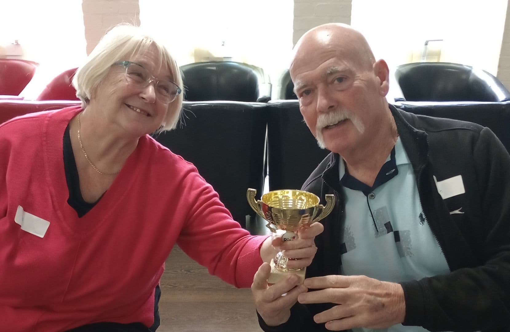 Two smiling older adults holding a gold trophy together while sitting indoors. - Home Instead