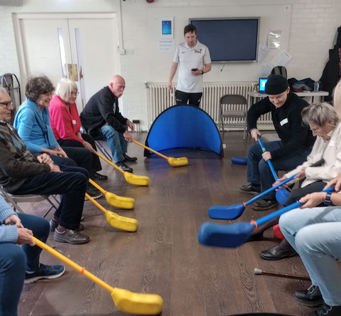 Older adults seated in a circle playing indoor hockey with foam sticks, guided by an instructor. - Home Instead