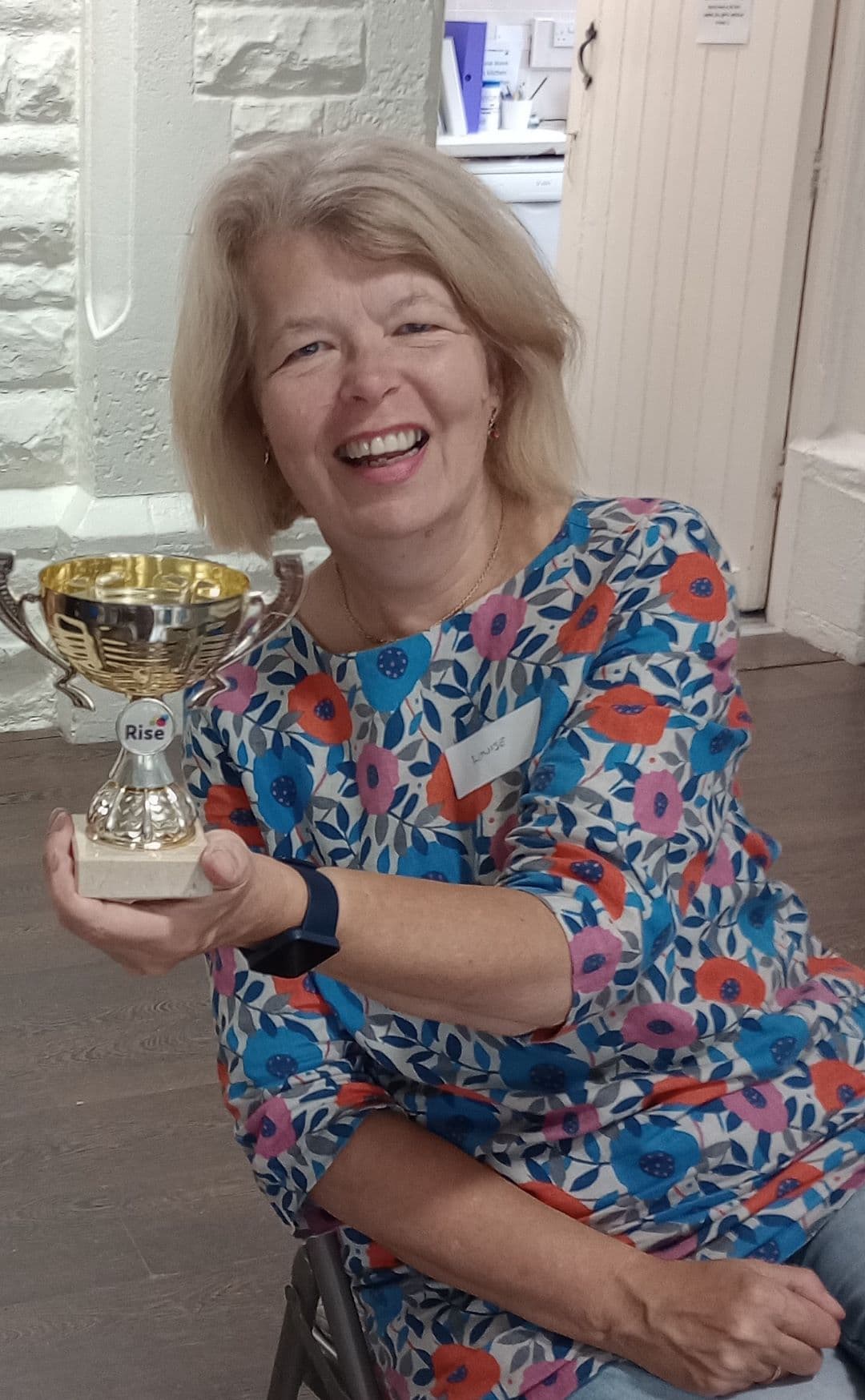 Smiling woman in a floral top holds a gold trophy while sitting indoors. - Home Instead
