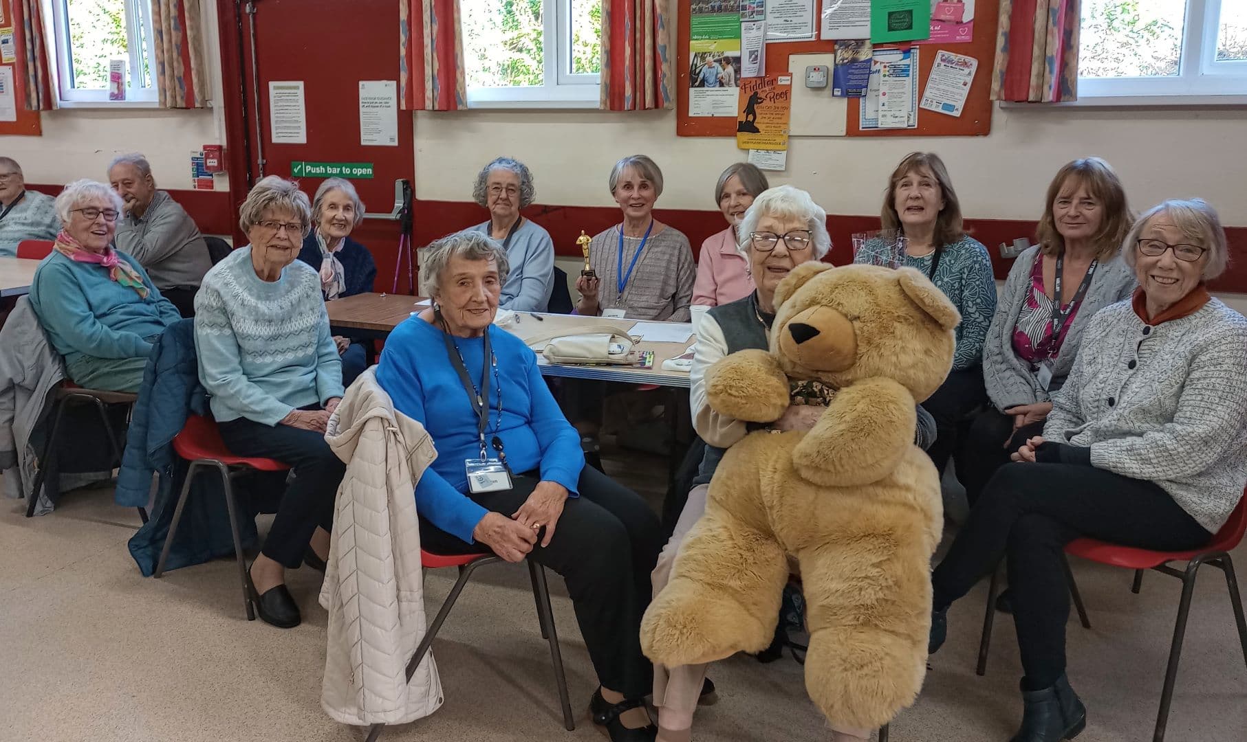 A group of smiling older women sit together indoors; one holds a large teddy bear. - Home Instead