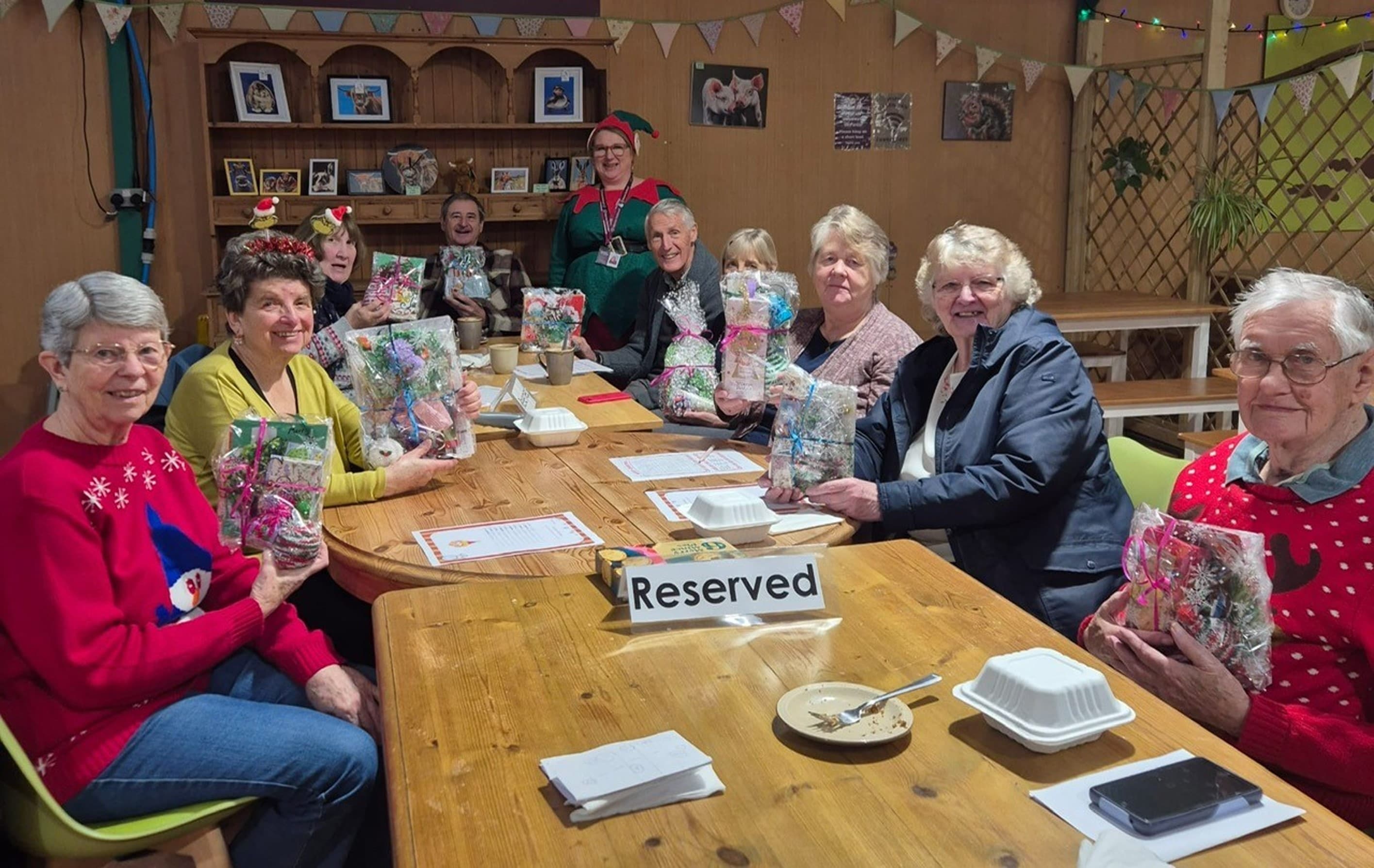 A group of smiling seniors holding gift bags sit around a table marked "Reserved" in a festive setting. - Home Instead
