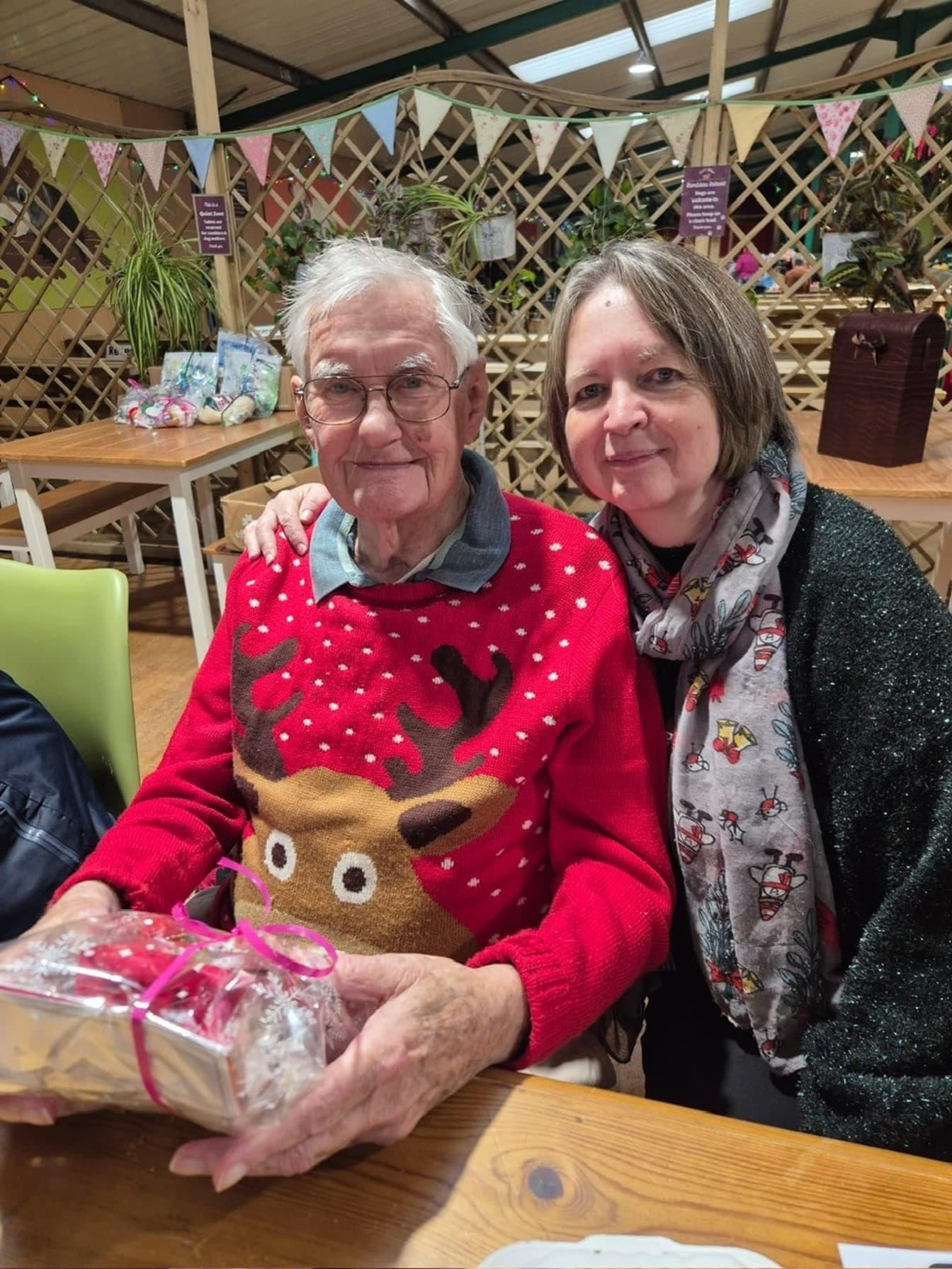 An elderly man in a reindeer sweater sits beside a smiling woman, holding a wrapped gift at a festive gathering. - Home Instead
