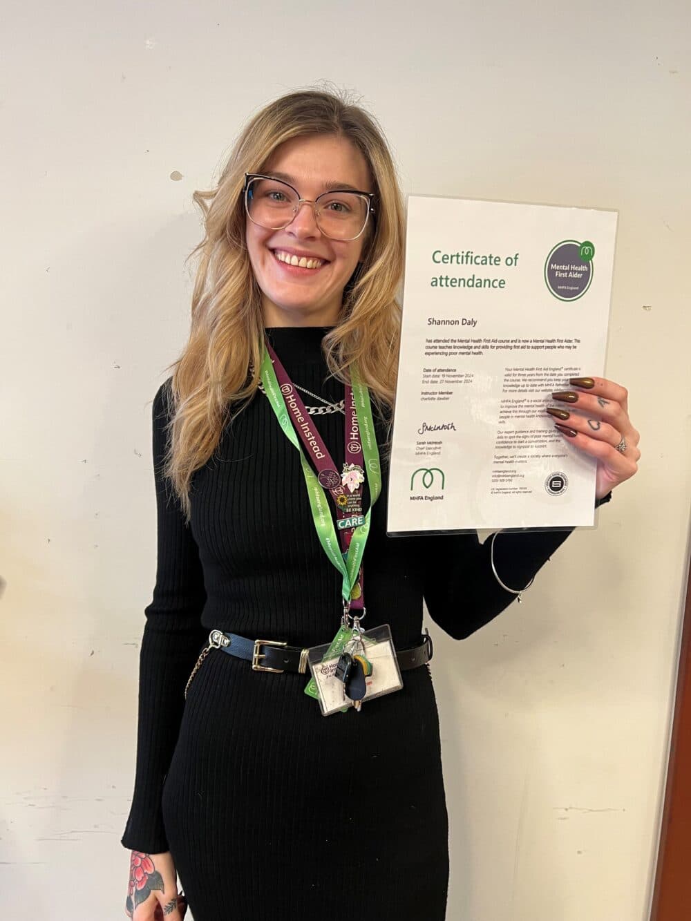 A smiling woman holds a certificate of attendance while wearing glasses and a black dress. - Home Instead