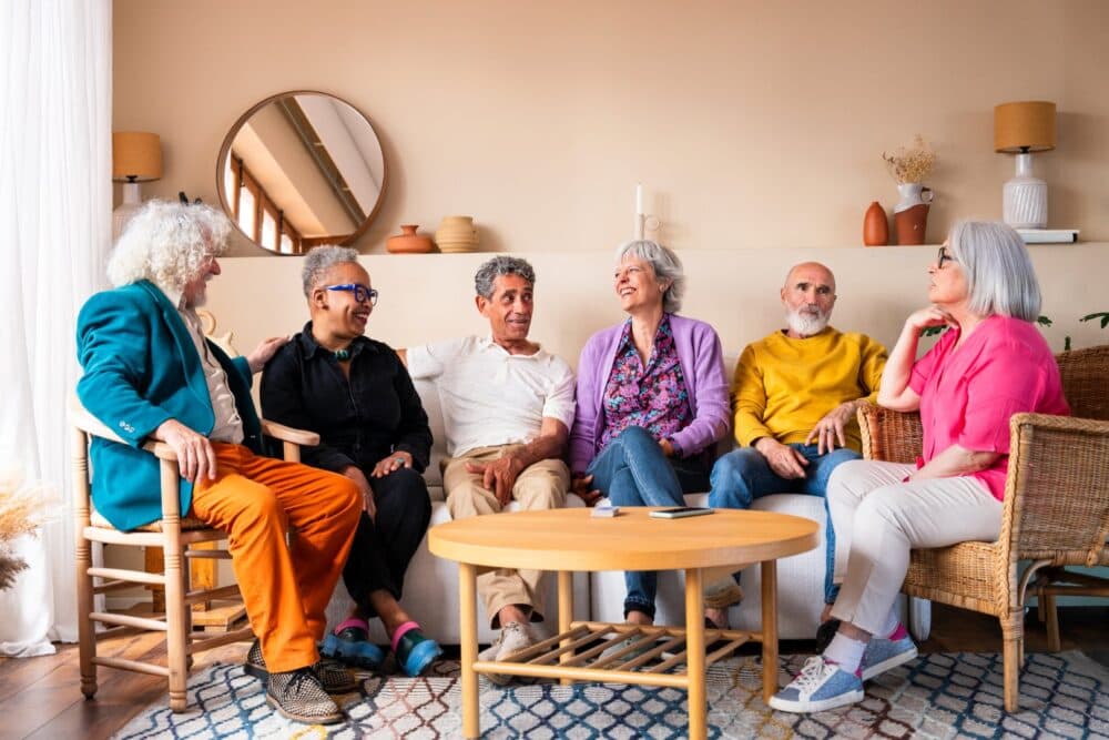 Six older adults sit together on a sofa, smiling and talking in a brightly lit living room. - Home Instead