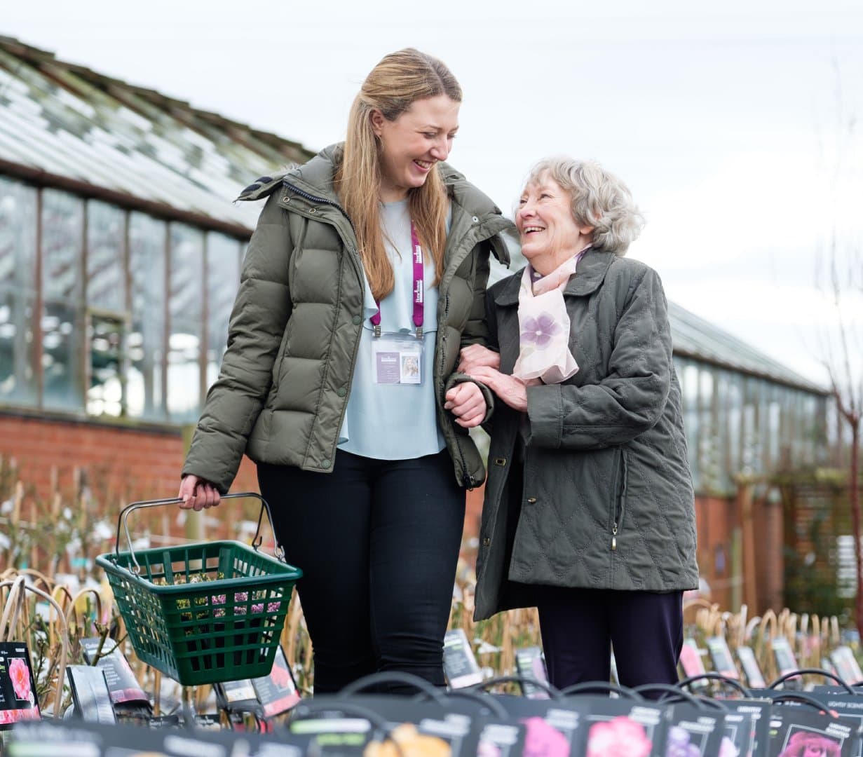 A young woman and an older woman smile together while shopping for plants outdoors. - Home Instead