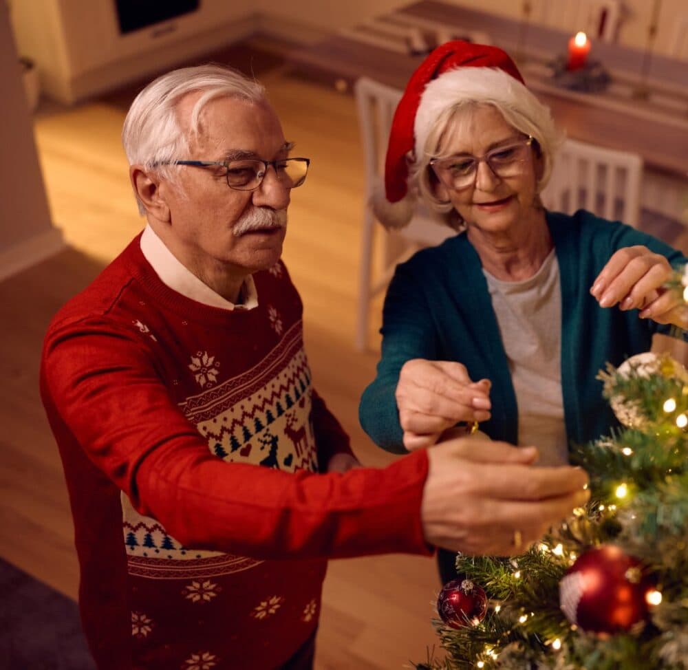 Two elderly people decorate a Christmas tree together, one wearing a Santa hat and the other a festive sweater. - Home Instead