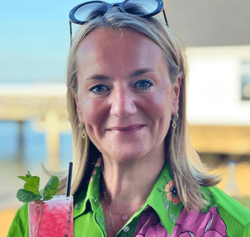 Smiling woman with sunglasses on her head holds a pink drink with mint, standing outdoors. - Home Instead