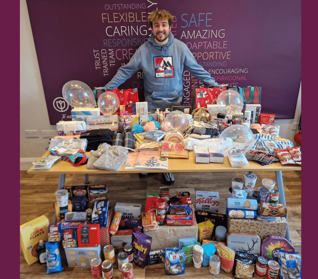 A person stands behind two tables filled with gifts, toiletries, food cans, and festive items. - Home Instead