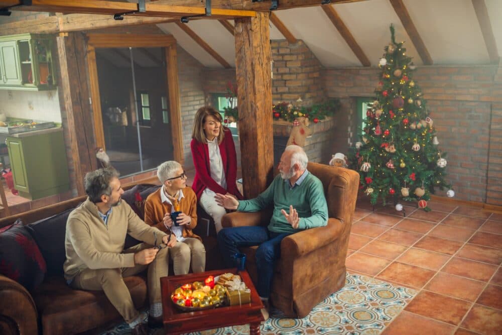 Four adults sit and talk in a cozy living room decorated with a Christmas tree and festive lights. - Home Instead