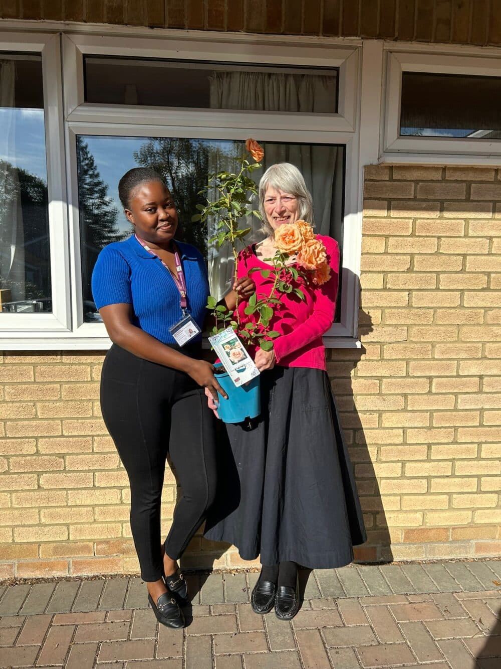 Two women standing outside a house, smiling, with one holding a potted rose plant in the sunlight. - Home Instead