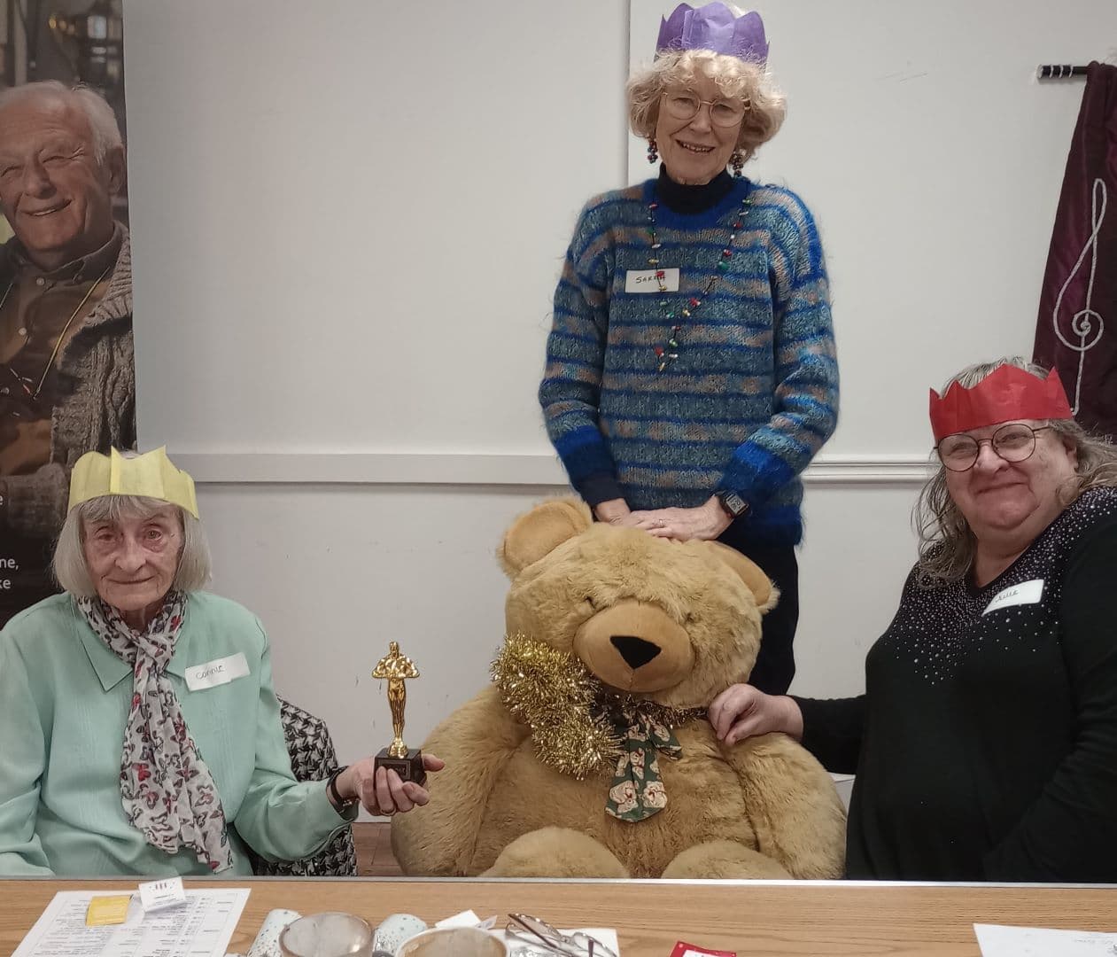 Three smiling women wearing paper crowns sit and stand with a large teddy bear at a decorated table. - Home Instead