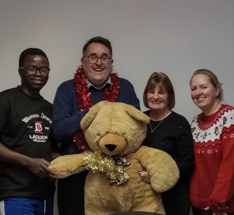 Four adults stand smiling indoors with a large stuffed teddy bear, wearing festive clothing and accessories. - Home Instead