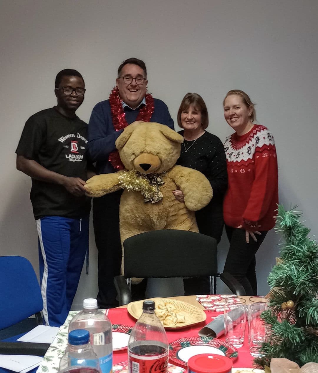 Four adults stand together indoors, smiling with a large teddy bear, festive decorations, and food on the table. - Home Instead