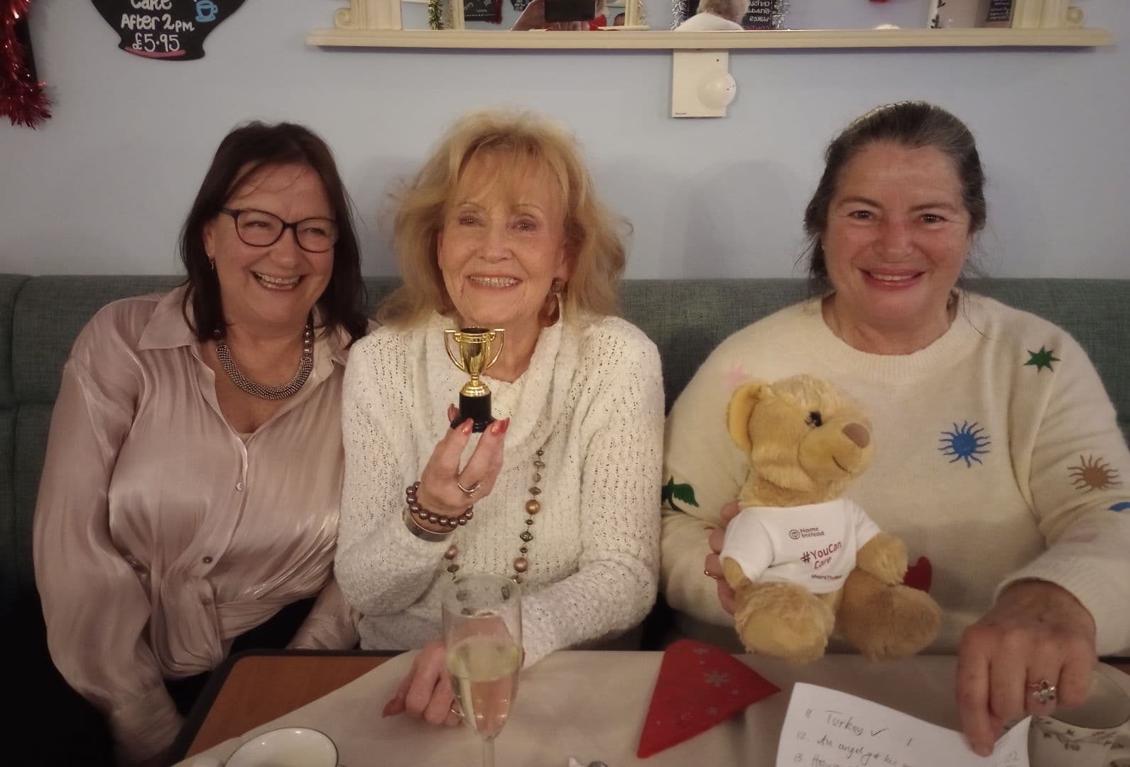 Three women smiling at a table; one holds a small trophy, another holds a teddy bear, drinks and paper nearby. - Home Instead