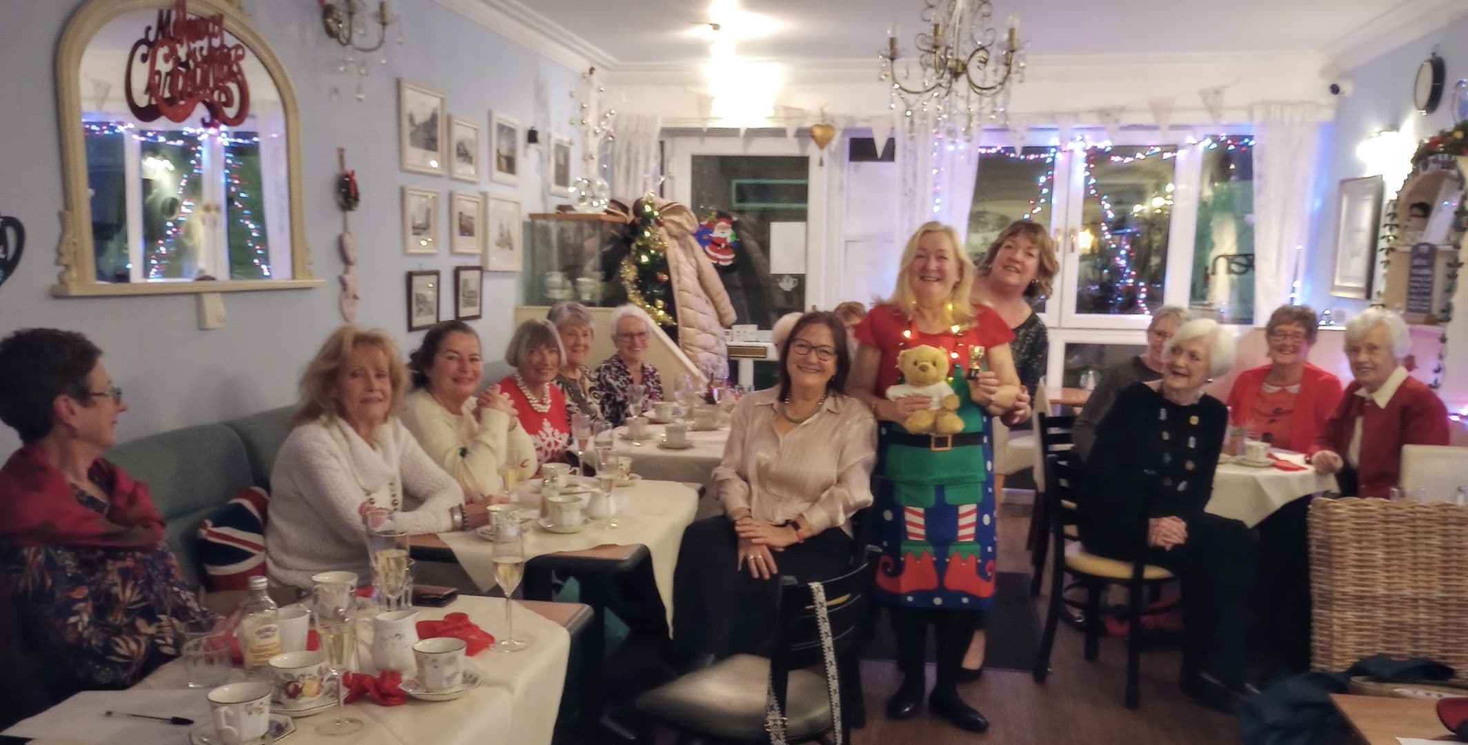 A group of smiling women sit around tables in a festive, Christmas-decorated room with tea and treats. - Home Instead