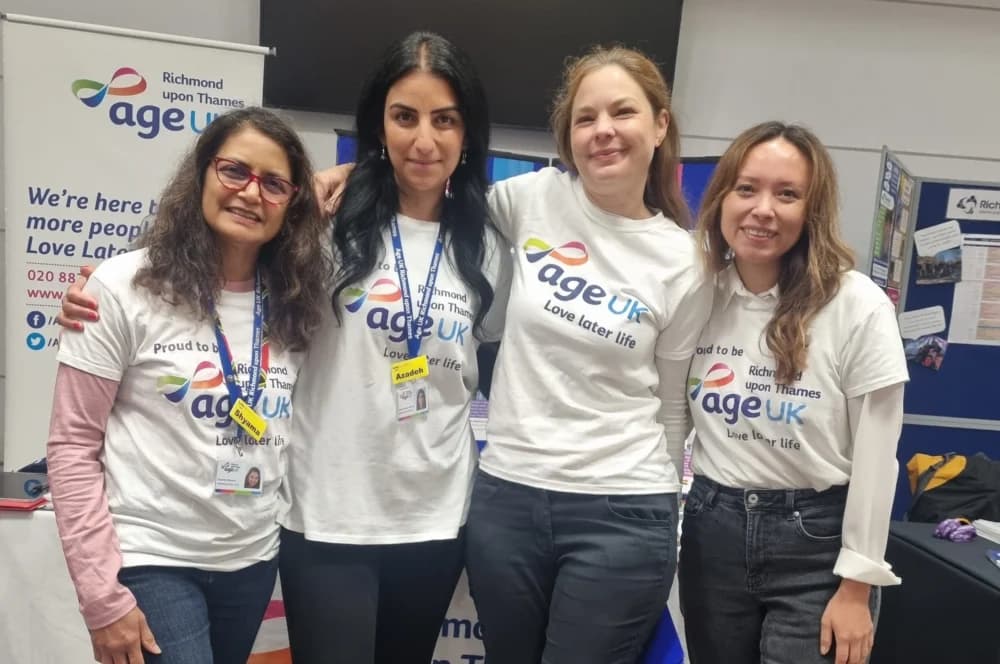 Four women smiling, wearing Age UK t-shirts, standing together at an informational event booth. - Home Instead
