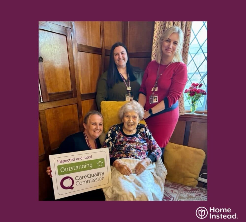 Two women at the back smiling and a senior woman sitting on the couch with another woman holding a CQC outstanding award sign, all smiling and happy