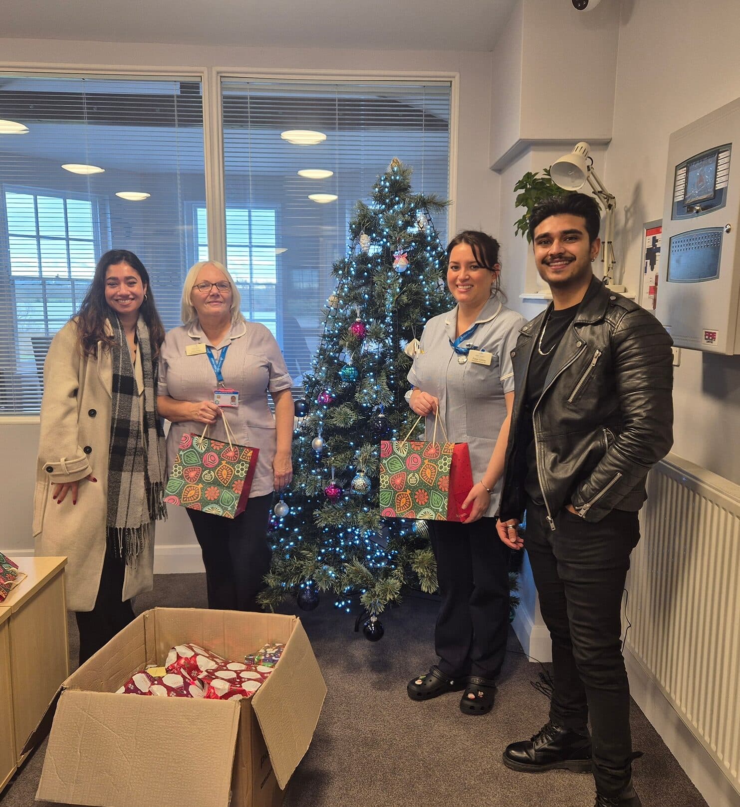 Four people stand by a Christmas tree holding gift bags, with a box of gifts on the floor nearby. - Home Instead