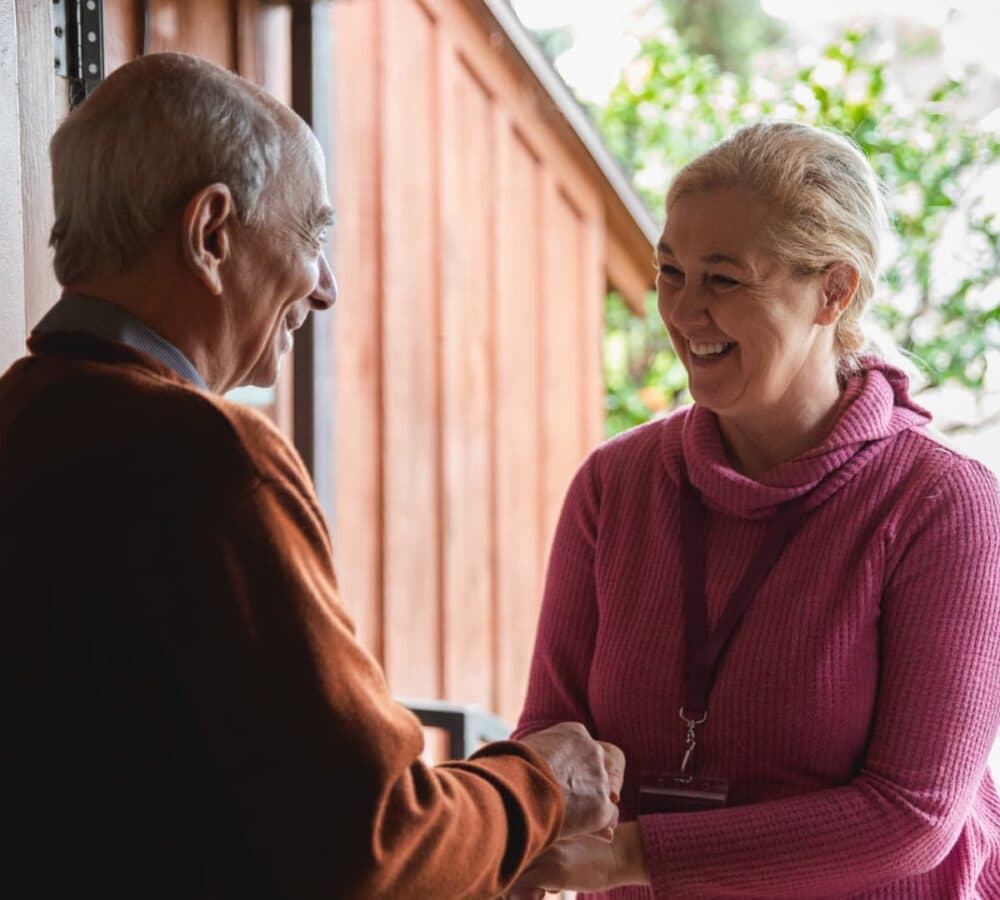 Lady carer wearing pink entering the door of the house while shaking the hand of a male senior