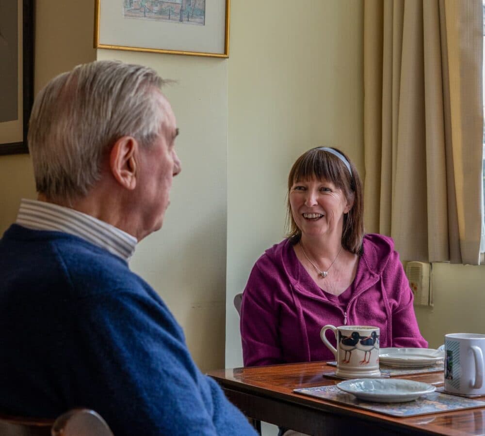 A senior man with grey hair and wearing blue sweatshirt sitting on a coffee table with tea cups and with his carer wearing purple and with short hair smiling