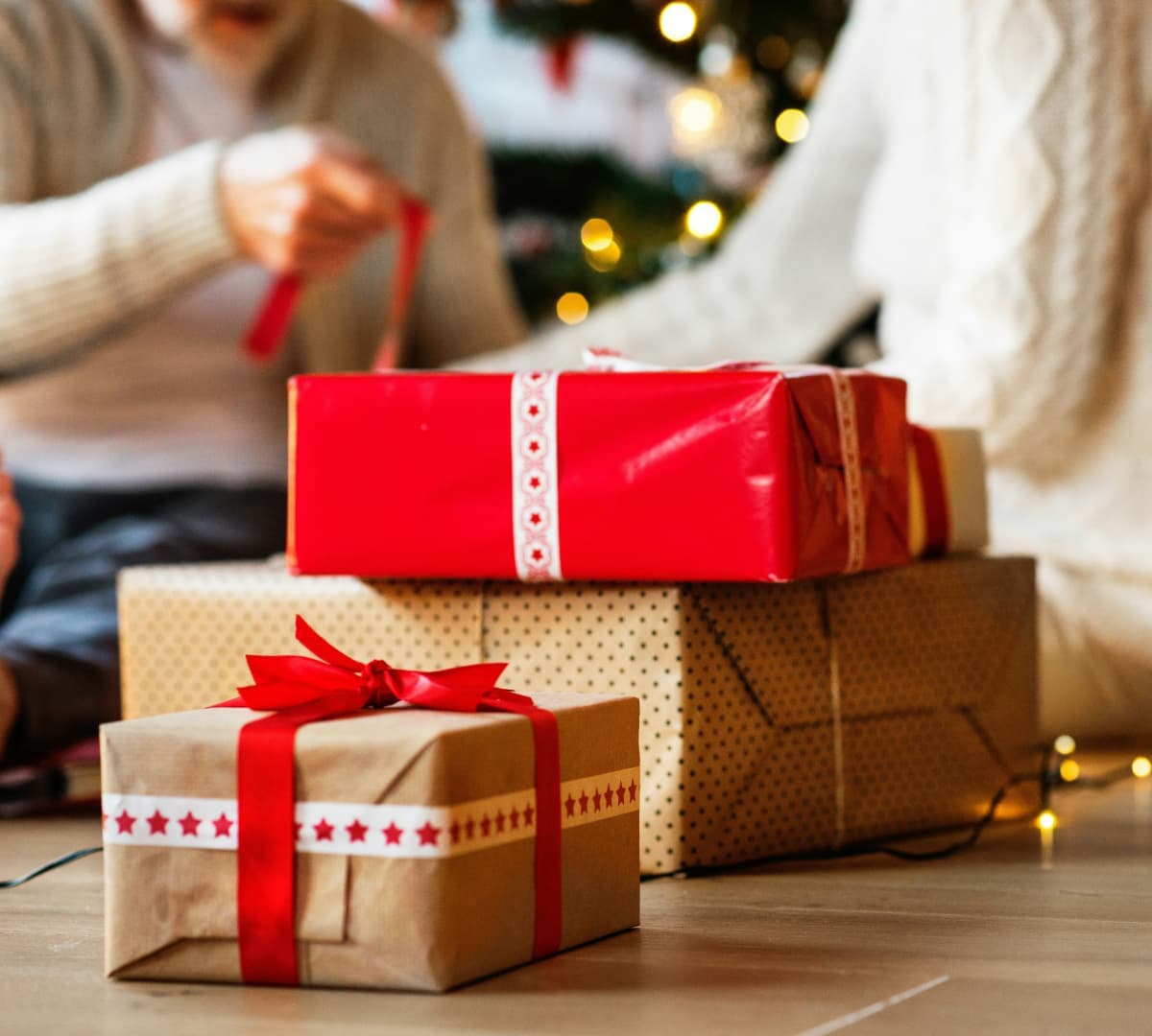 Three Christmas gifts on the floor in brown and red wrapper with two people wrapping at the background
