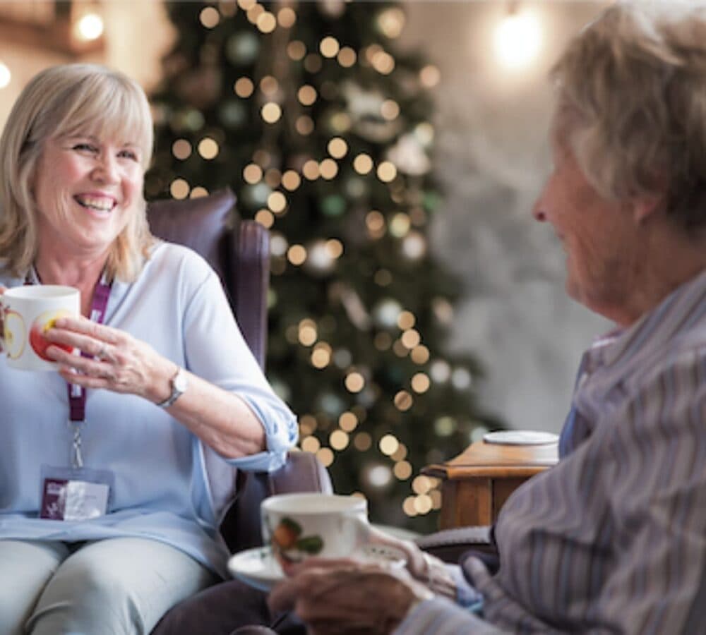 Senior woman sitting down and drinking coffee with her carer smiling and happy with Christmas tree at the background