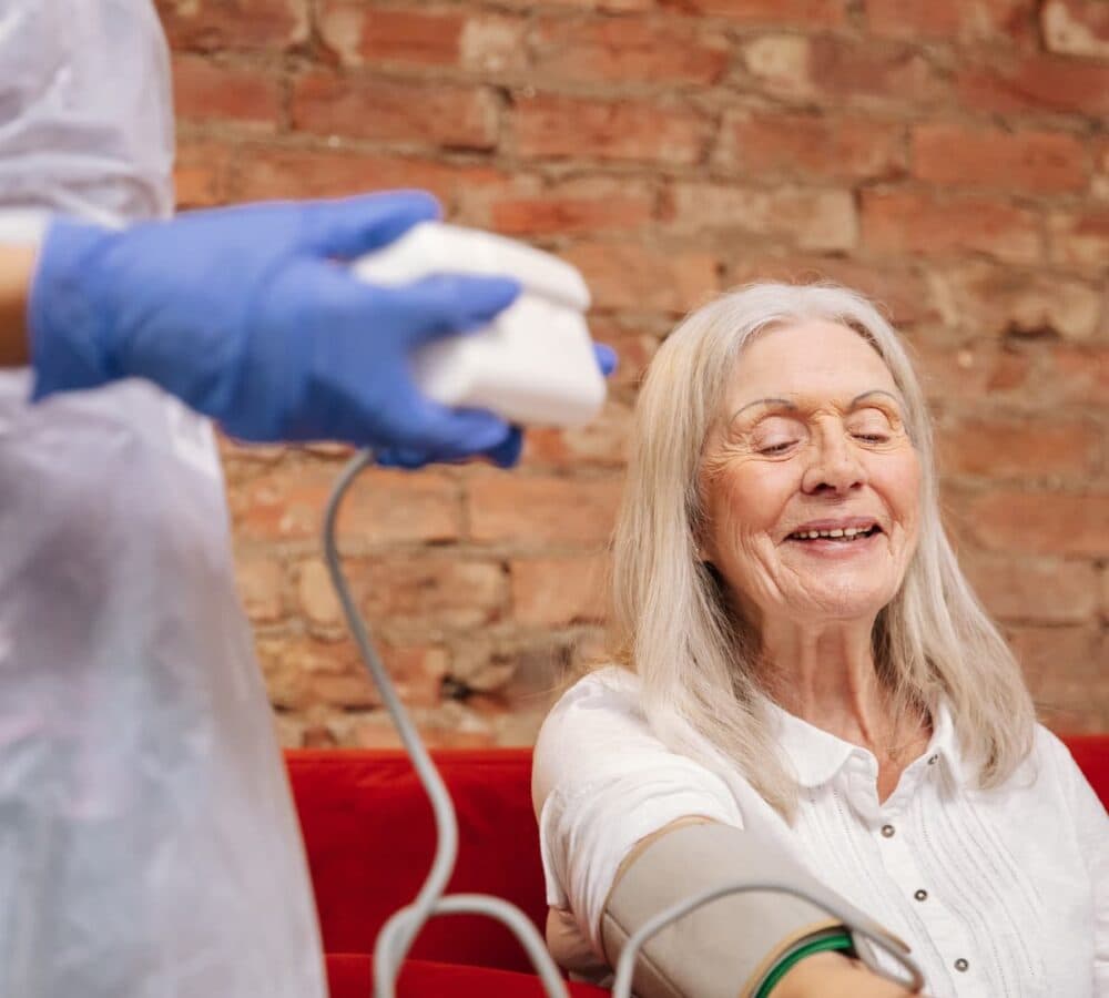 Elderly woman with long white hair blood pressure being taken by her carer wearing blue gloves