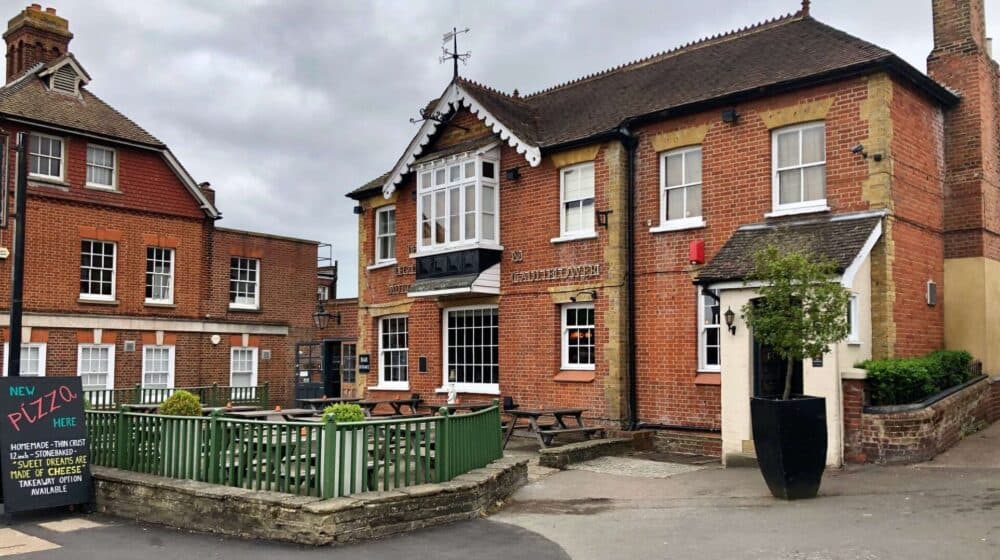A red-brick pub with a patio, outdoor seating, and a sign advertising pizza under a cloudy sky. - Home Instead