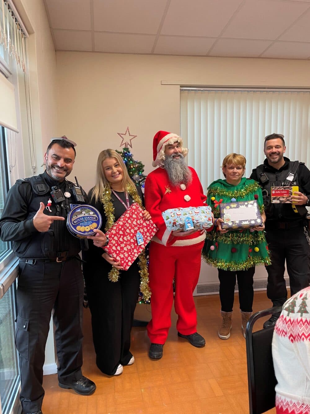 Five people, including two police officers, Santa, and two women with gifts, stand by a Christmas tree indoors. - Home Instead