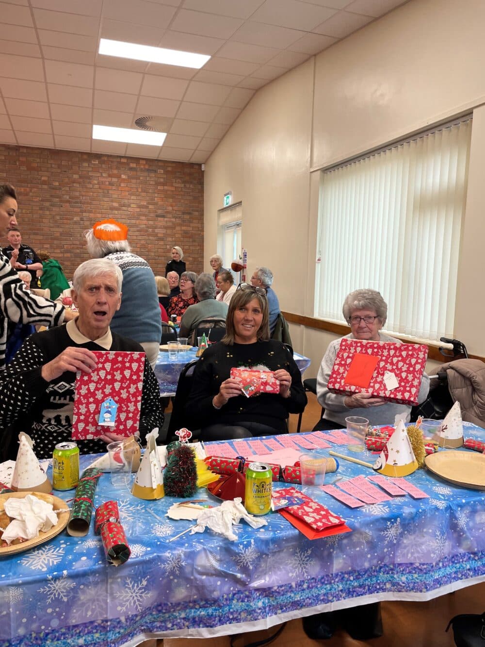 Three people at a festive table hold wrapped gifts at a holiday party in a community hall. - Home Instead