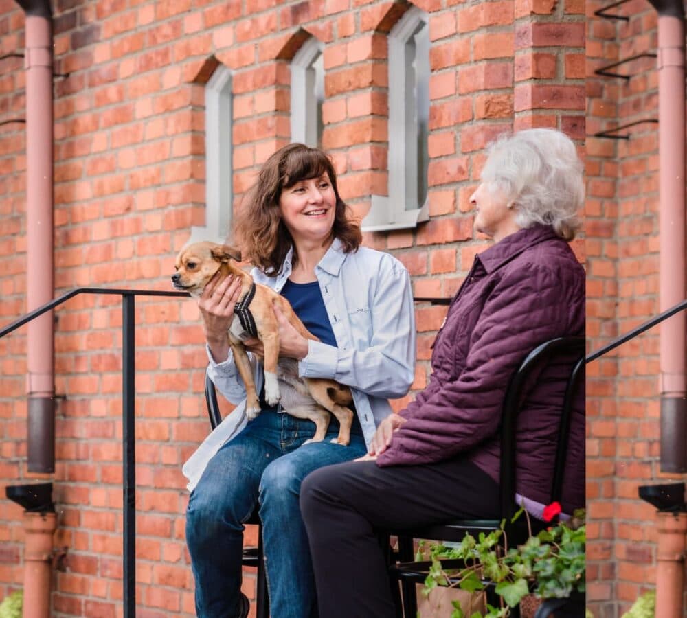Woman holding a dog while sitting outside the house smiling and chatting with her mother