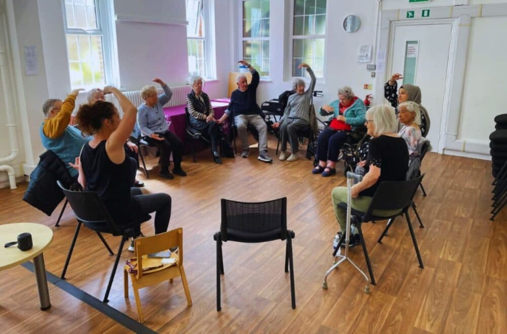 A group of seniors sit in a circle doing seated arm exercises in a bright room. - Home Instead
