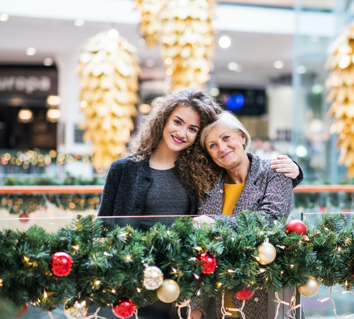 Senior woman with her daughter inside a mall both happy and smiling with Christmas decors and lights in front and at their back