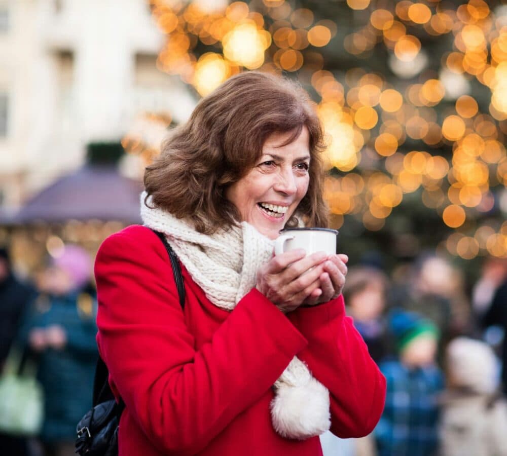 Older adult sipping coffee outdoor while smiling and happy with Christmas lights at the back