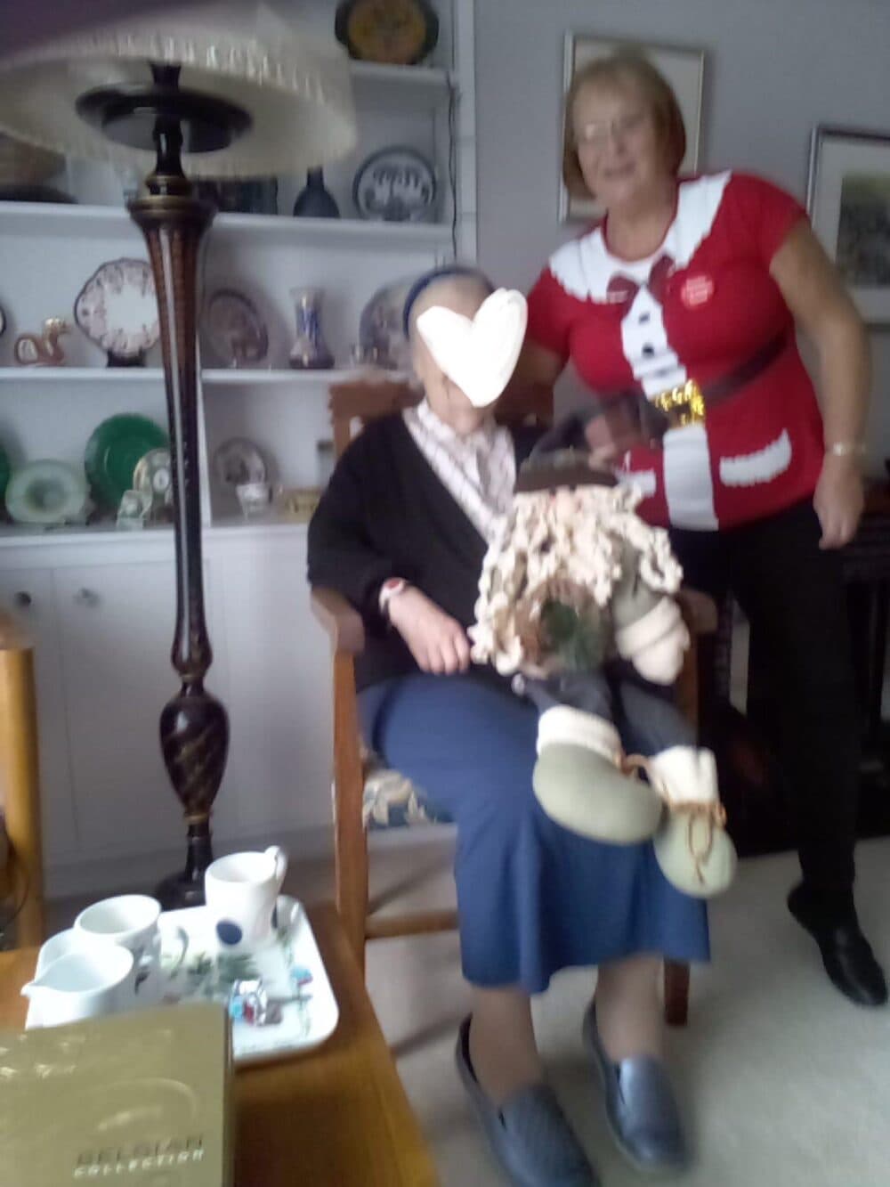 Two women, one seated and one standing, in a living room with shelves of dishes and a tea set on a table. - Home Instead