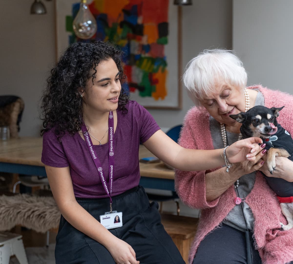 Senior woman with white hair happy while holding her black dog inside her home with her carer wearing purple and with curly hair both sitting down on a couch