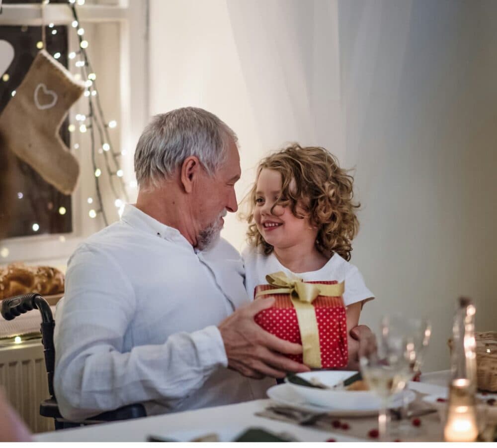 Senior man with grey hair sitting on a chair while holding a gift from a girl with curly hair